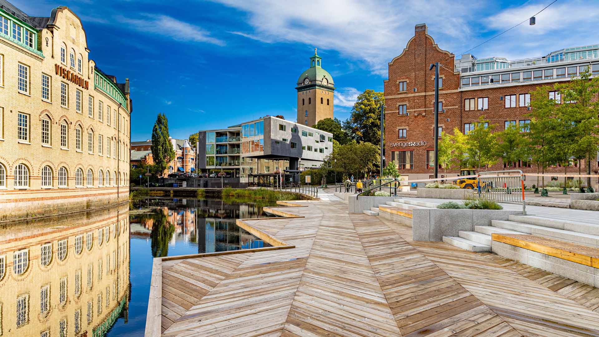 En stadsvy med en träbrygga vid en å och siluetten av en kyrka och stad i bakgrunden