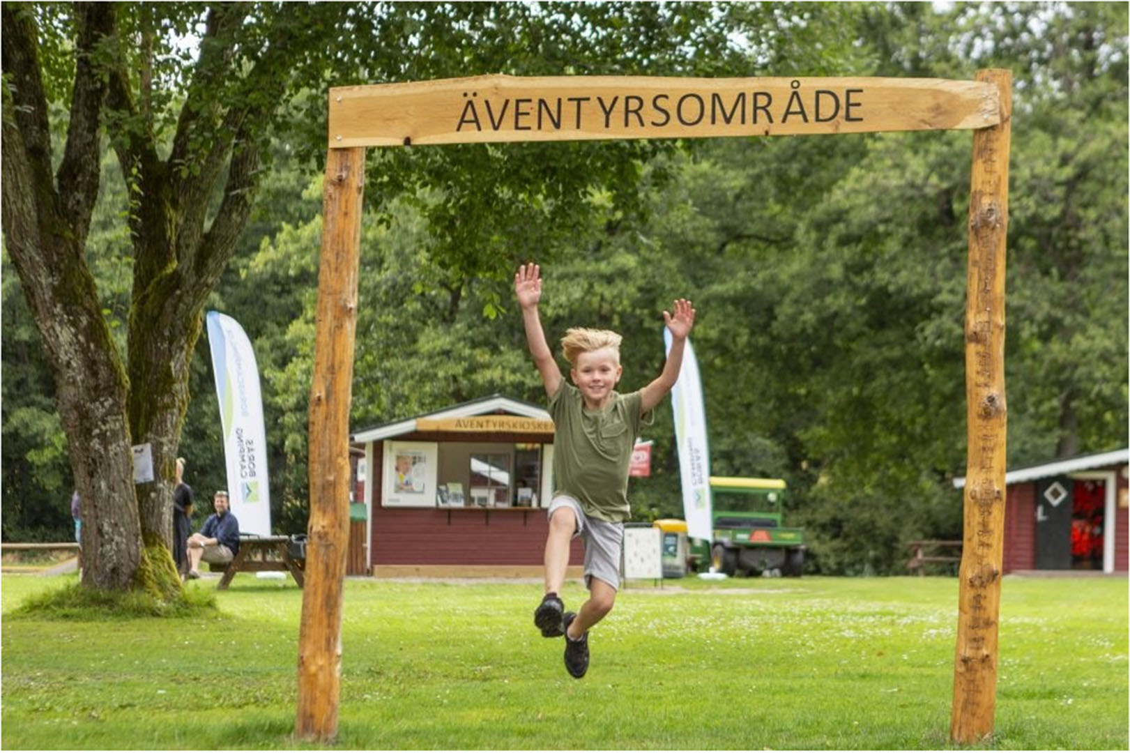 Stor träport/skylt som talar om att det är ett äventyrsområde. En liten kille, ca fem år hoppar och sträcker upp armarna under porten.